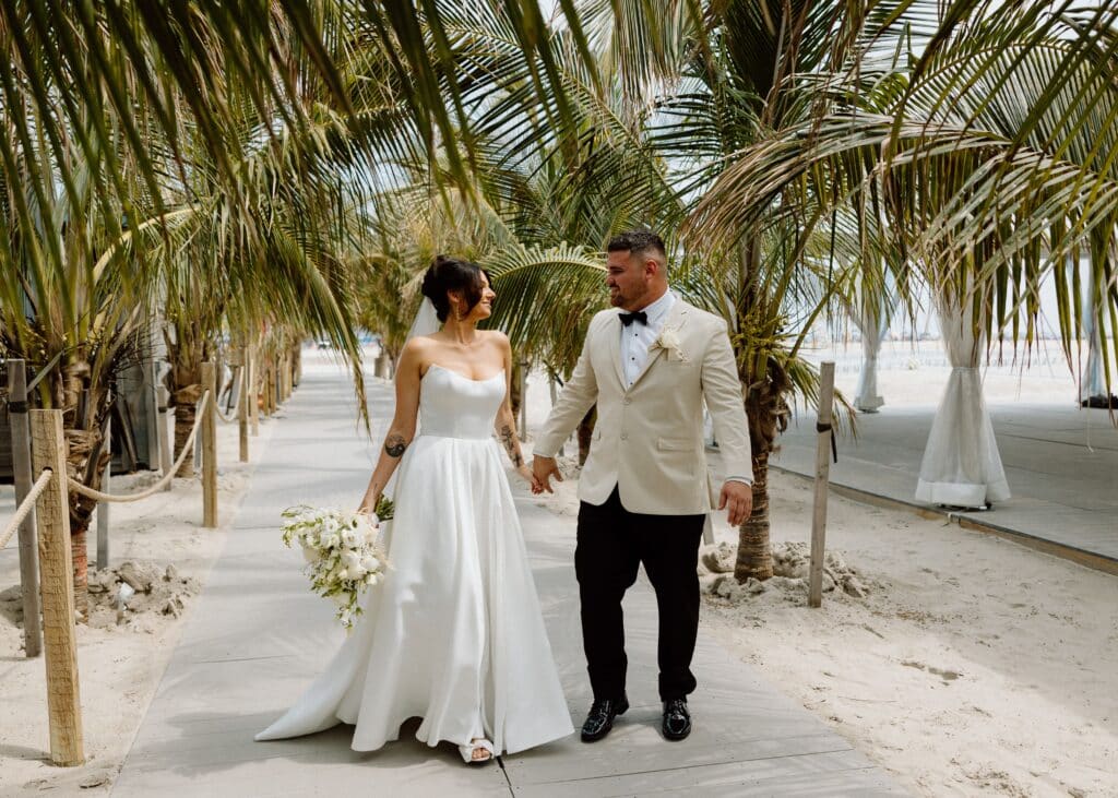 Bride and groom holding hands between palm trees at ICONA Diamond Beach wedding venue