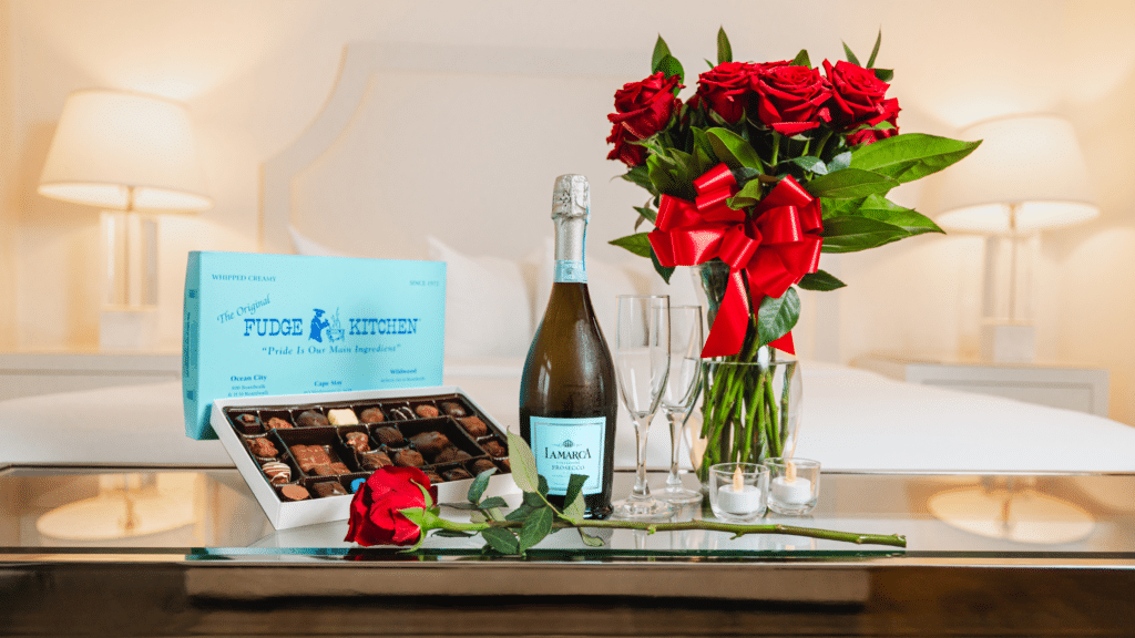 A box of chocolates, bouquet of roses, and a bottle of champagne on a table in front of an ICONA Resorts hotel room bed